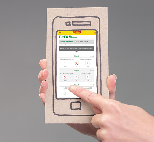 Finger zeigt auf ein Handy mit TOTO Ergebniswette Spielschein